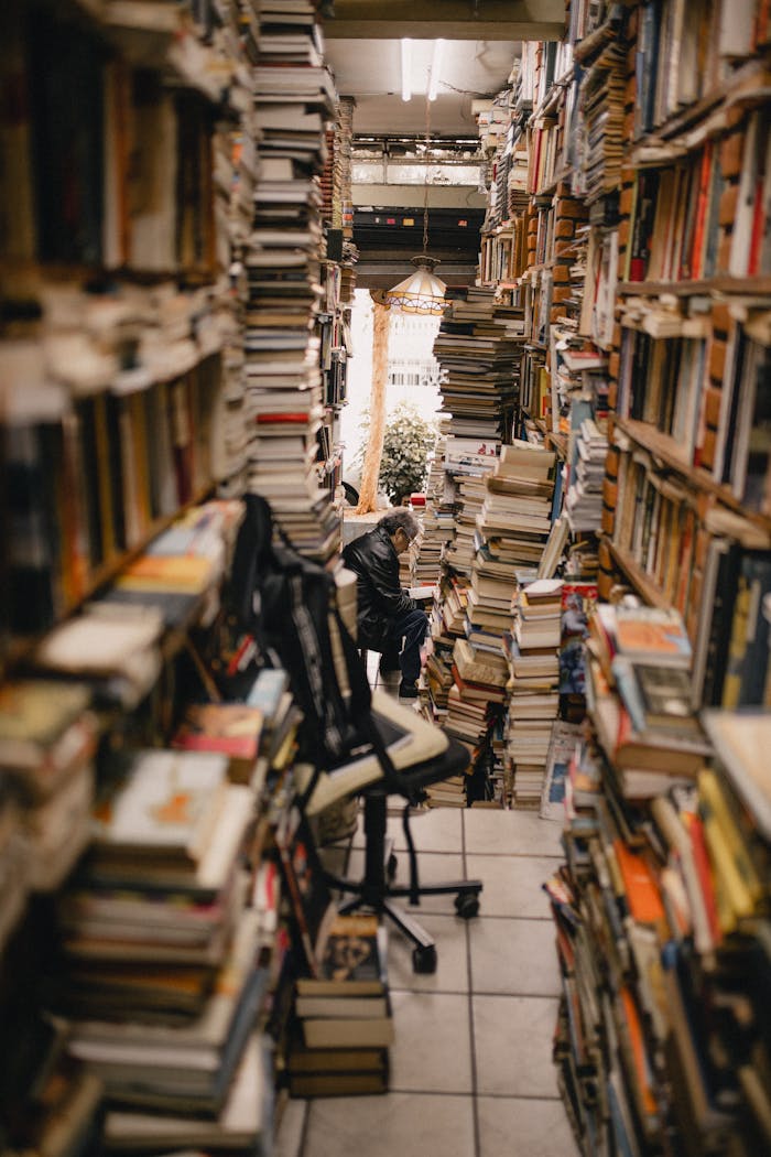 A narrow aisle filled with stacks of books in a cozy, crowded bookstore, featuring a person reading.